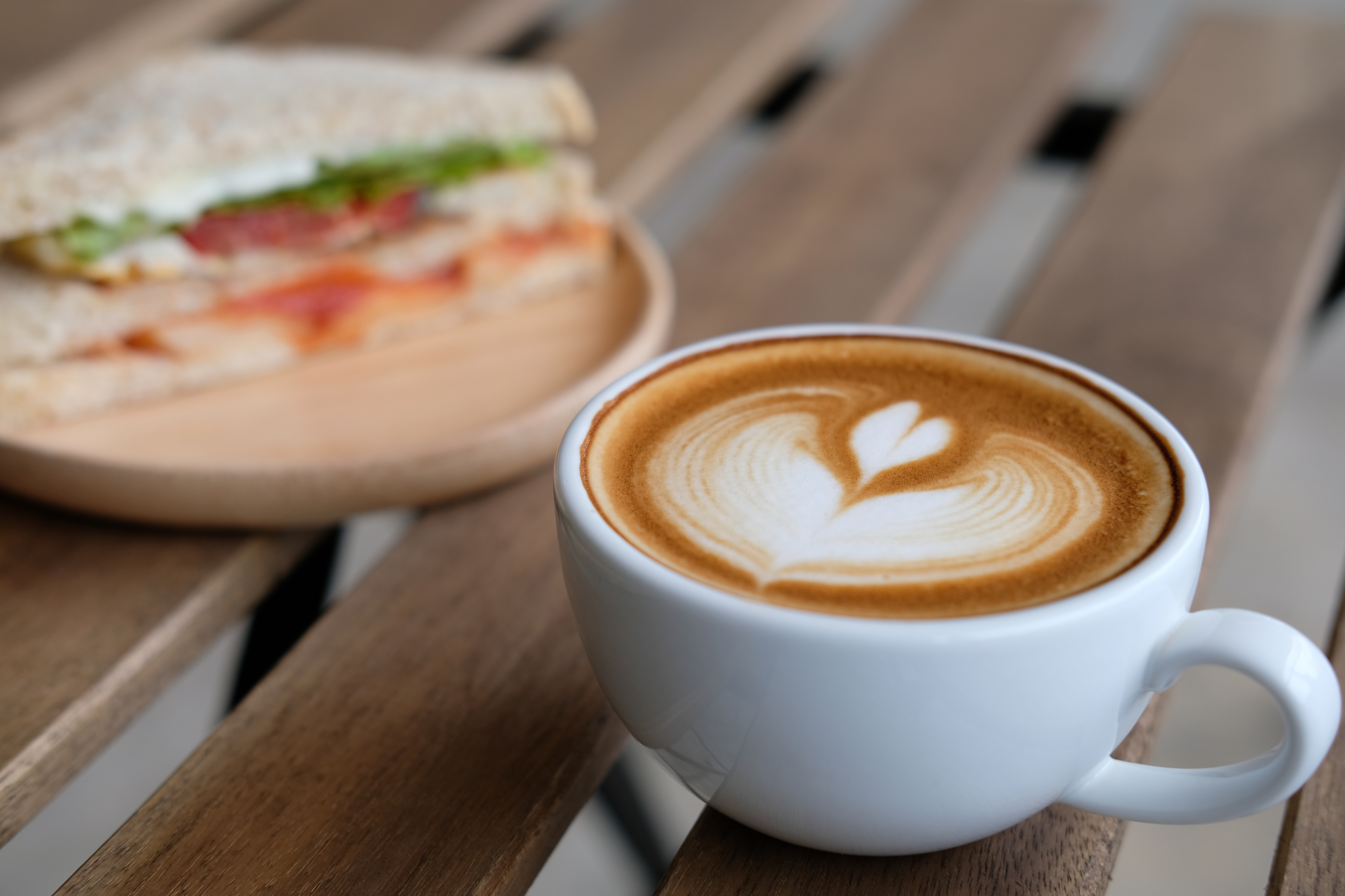 A latte in a white mug on a wooden table with a sandwich in the background. 