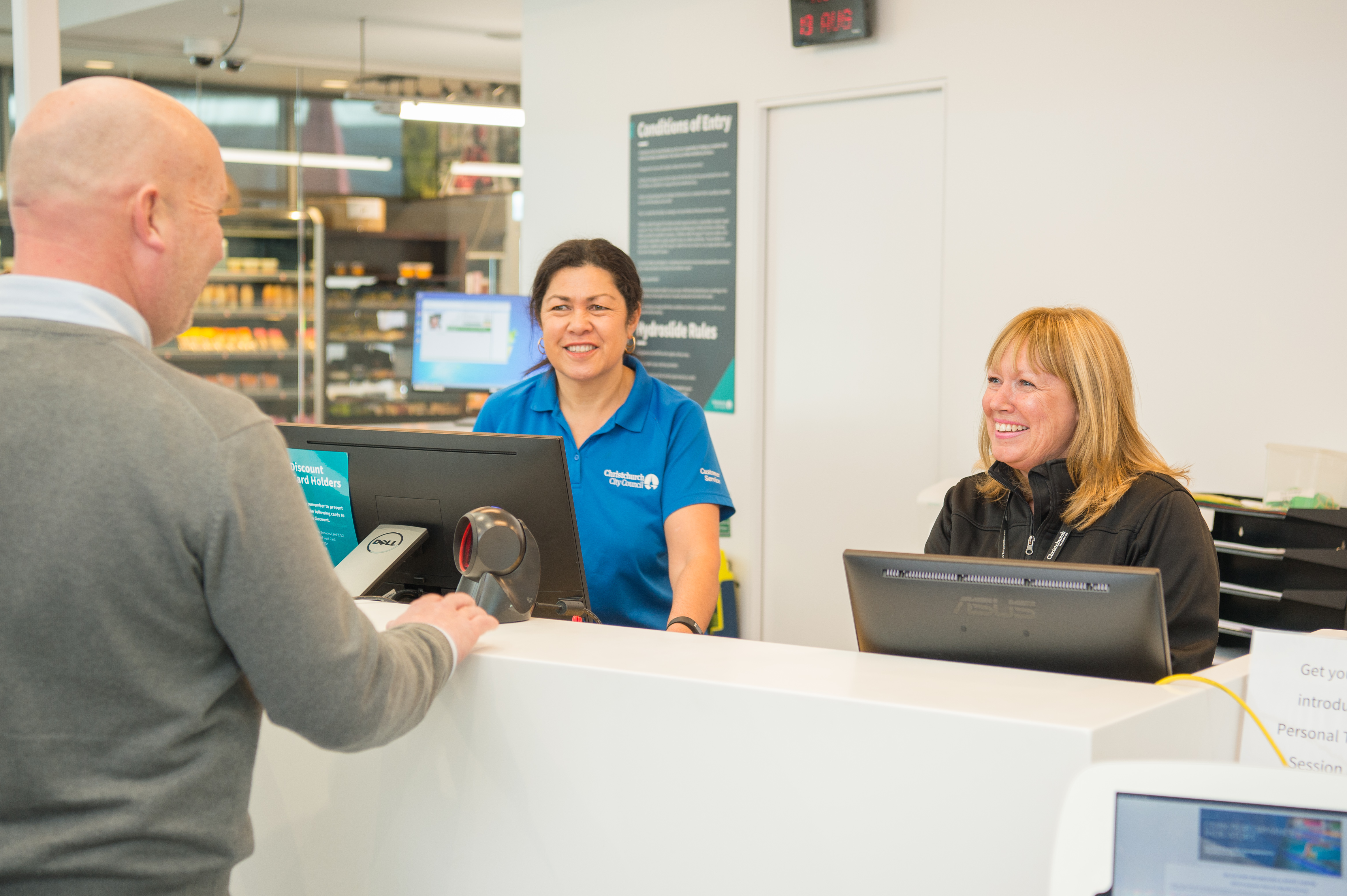 Two smiling women staff members standing behind the counter at Jellie Park Rec Centre talking to a man in a grey sweater. 