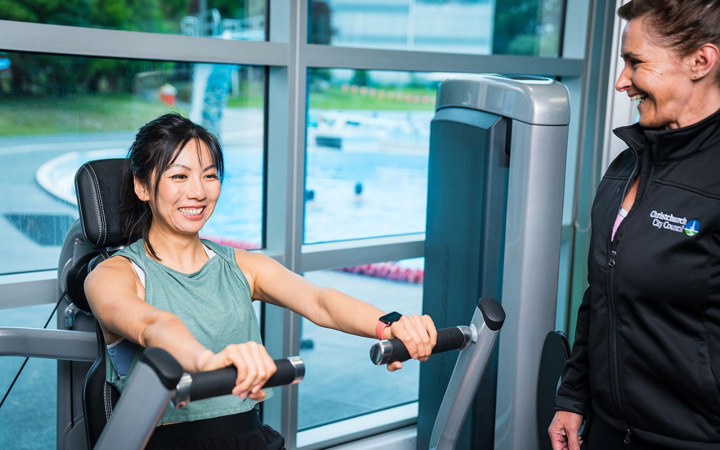 A female personal trainer in a black staff uniform assisting a female client with a shoulder press at Christchurch Recreation and Sport Centre.