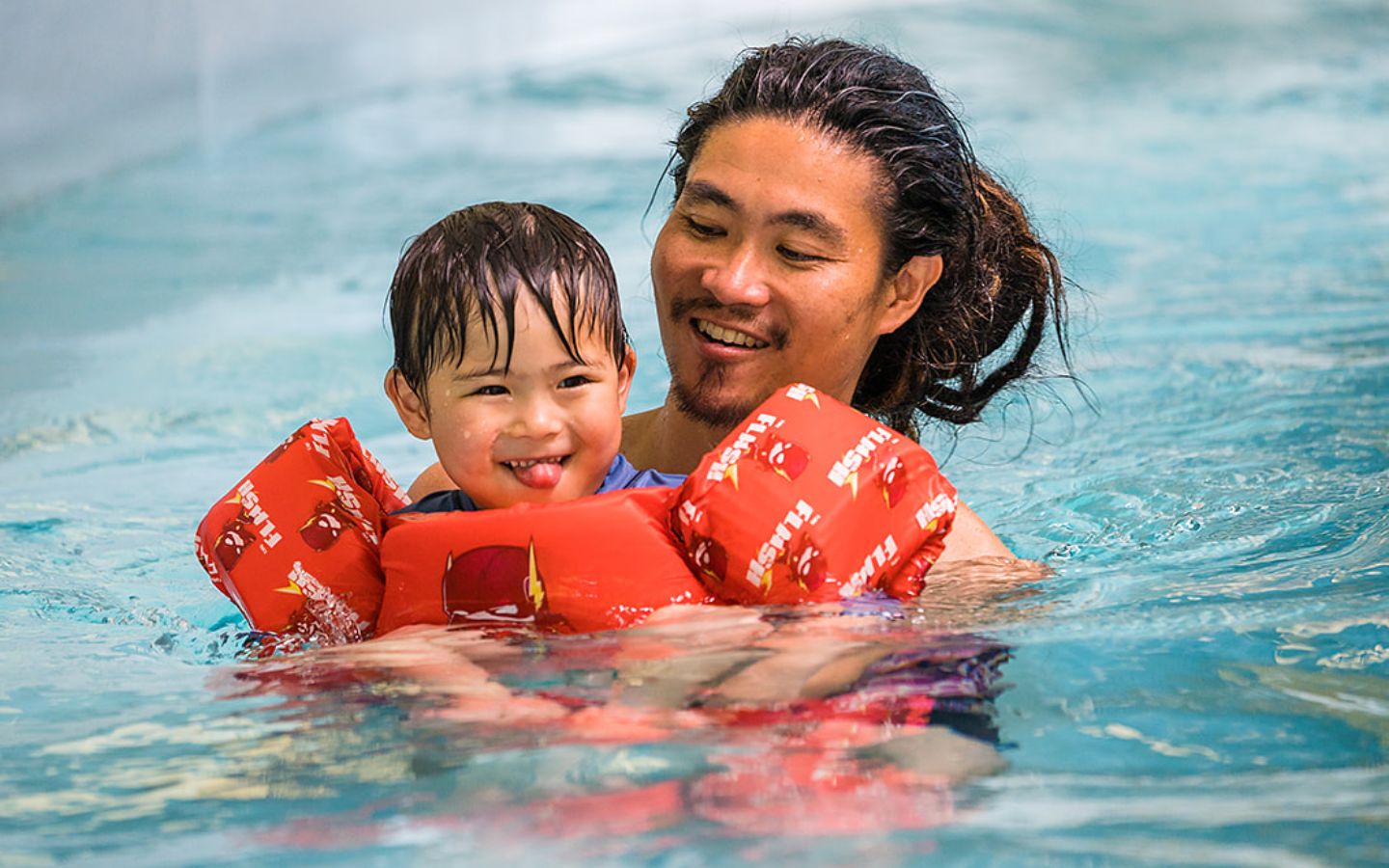 A little boy with a red life jacket is being held by a smiling man in Christchurch Recreation and Sport pool. 