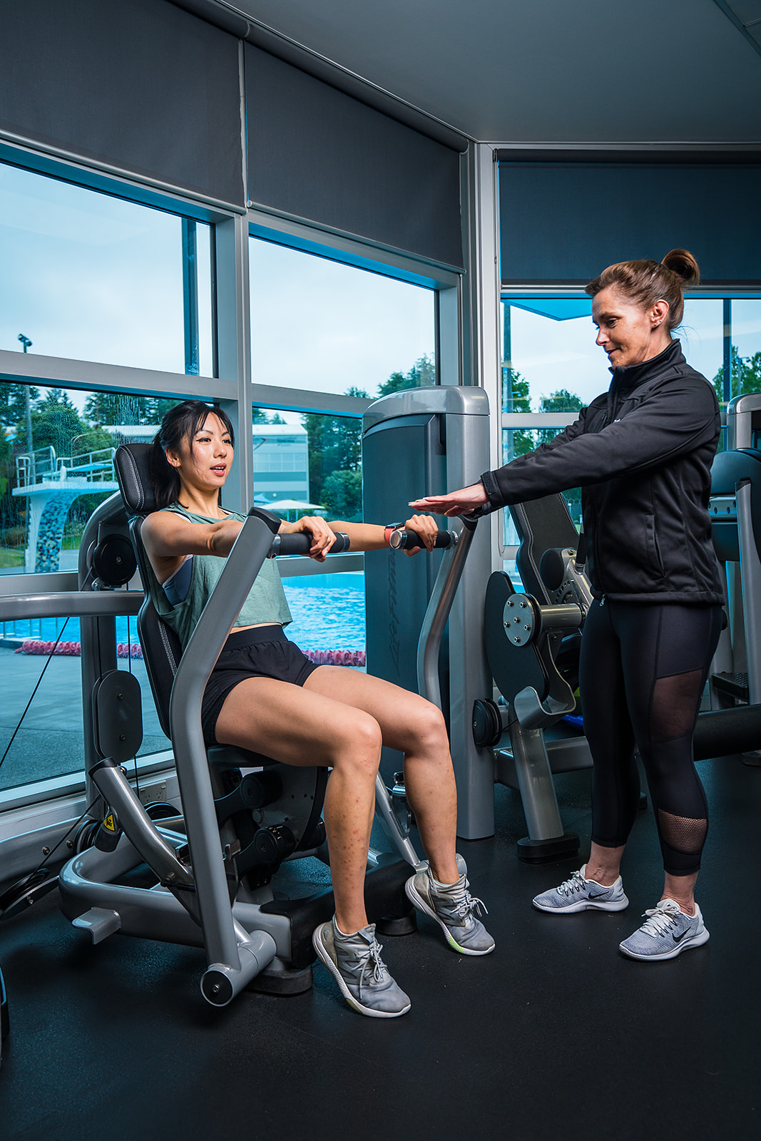 A female personal trainer in a black staff uniform assisting a female client with a shoulder press at Christchurch Recreation and Sport Centre.
