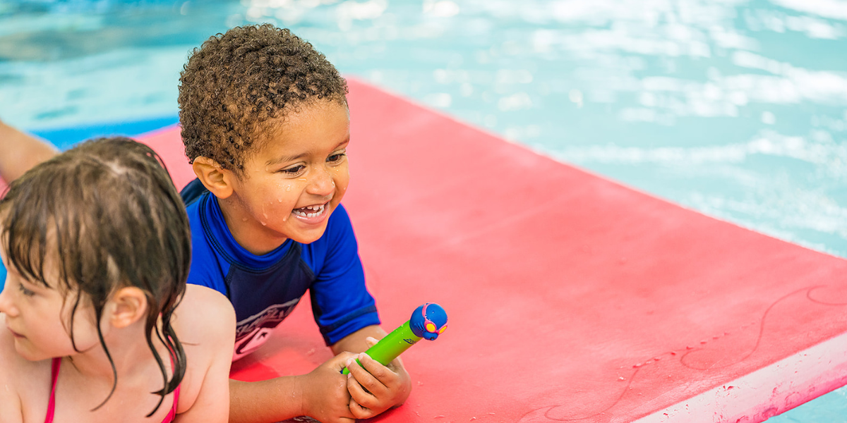 A little boy and girl laying on a red flotation carpet at Christchurch Recreation and Sport pool. 