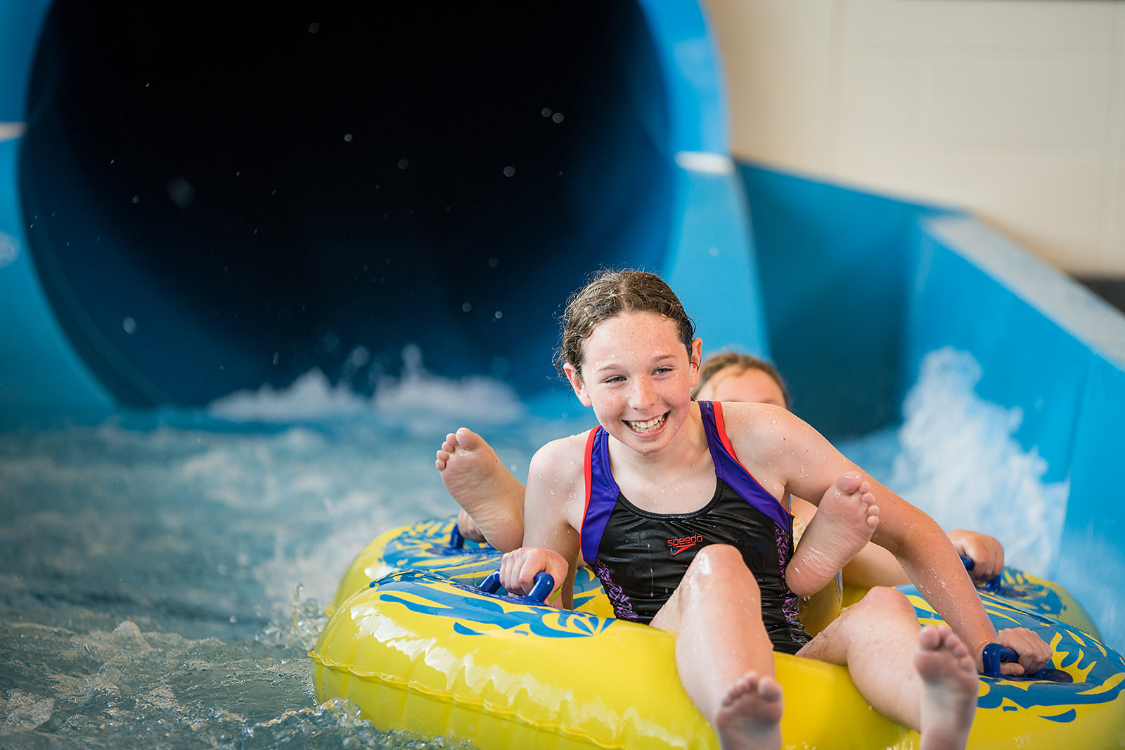 Two smiling girls sliding down the blue hydroslide in a yellow raft at Christchurch Recreation and Sport pool.