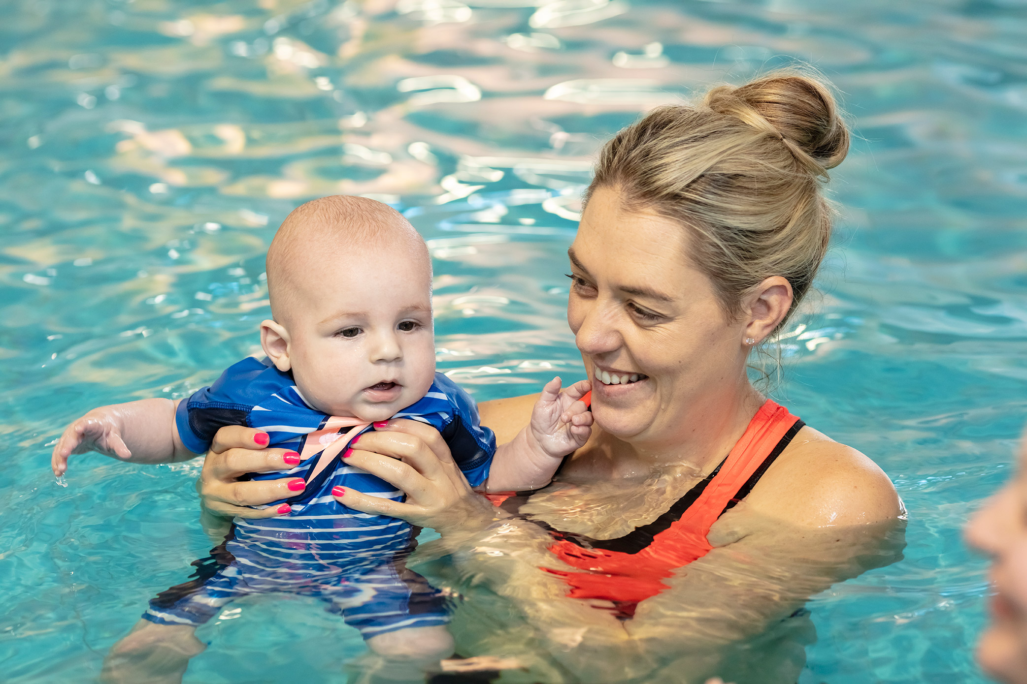 A smiling woman holding onto a baby while attending a Tiny Turtles swim class at Pioneer Recreation and Sport Centre. 
