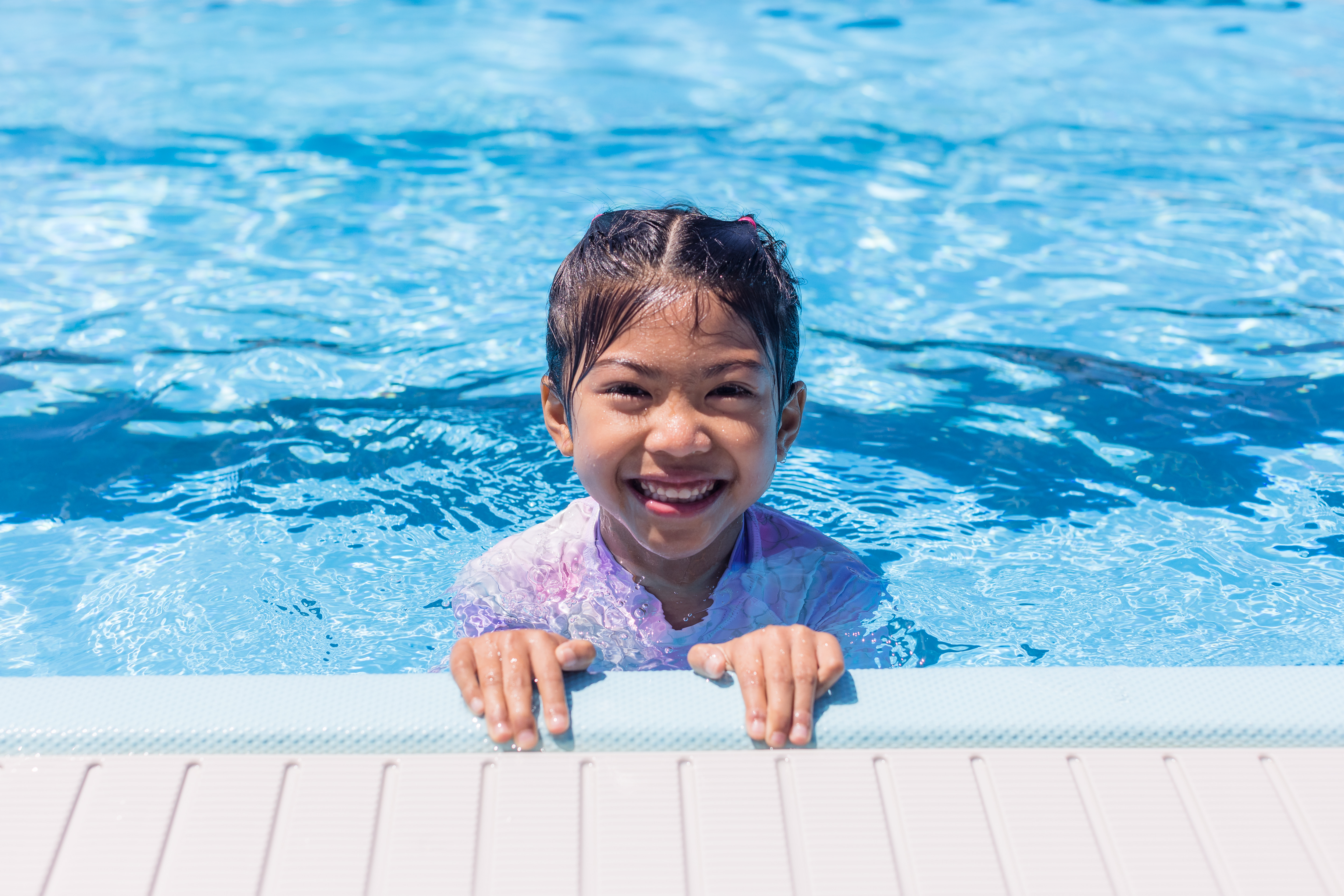 A smiling little girl wearing pink swimmers playing in the toddler pool at the Halswell Summer pool. 