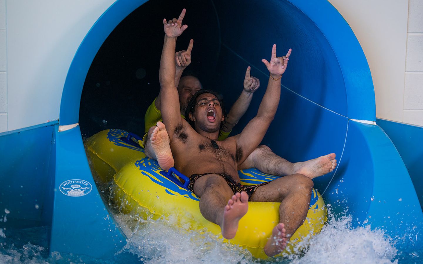 Two smiling men sliding down the blue hydroslide at QEll Recreation and Sport Centre in a yellow tandem raft. 