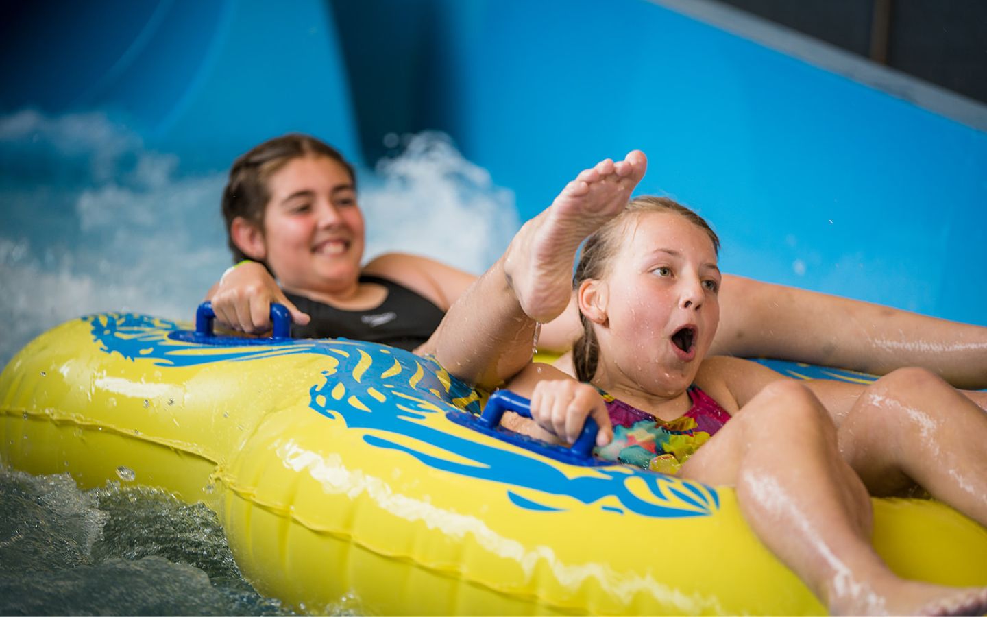 Two smiling girls sliding down the blue hydroslide at QEll Recreation and Sport Centre in a yellow tandem raft. 