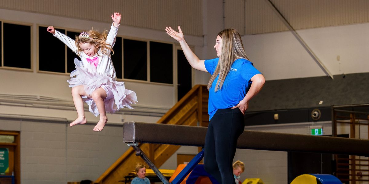 A little girl jumping off a balance beam with the help of an instructor at Christchurch Recreation and Sport centre. 