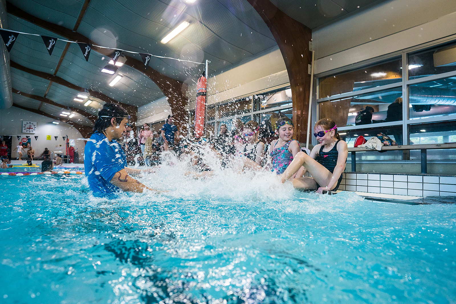 A male swim instructor taking to a row of children who are seated on the pool deck splashing at Christchurch Recreation and Sport pool. 