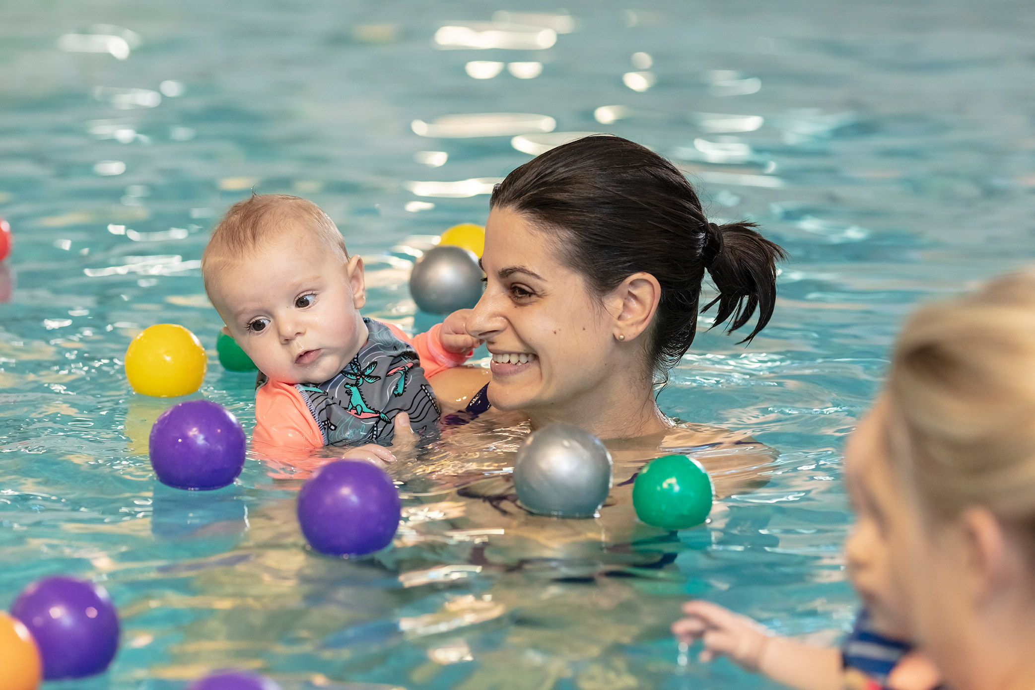 A toddler surrounded by floating balls while being held by a smiling woman in a Christchurch Recreation and Sport pool. 