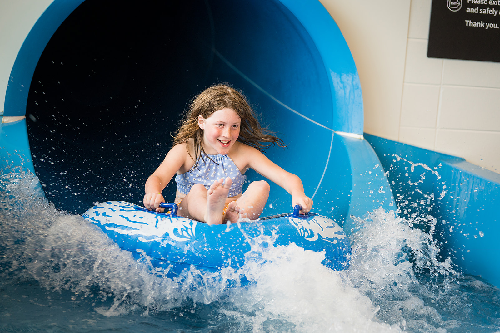 A smiling girl sliding down the blue hydroslide in a blue raft at Christchurch Recreation and Sport pool.
