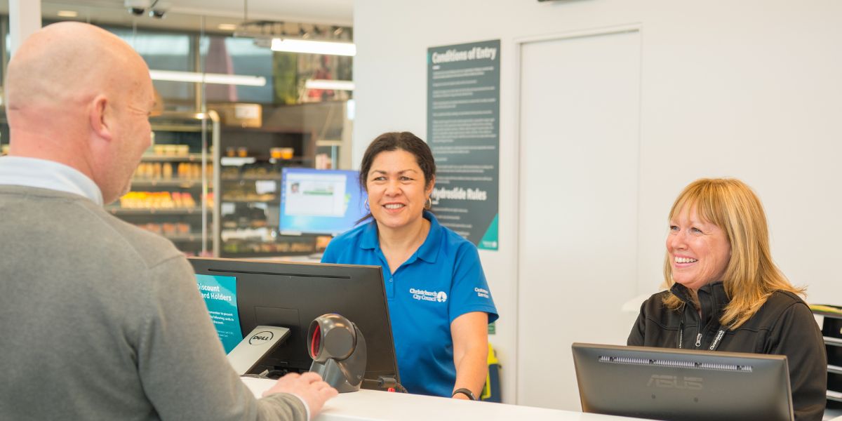 Two smiling women staff members standing behind the counter at Jellie Park Rec Centre talking to a man in a grey sweater. 