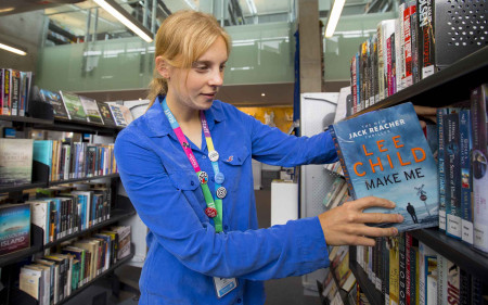 A library staff member in a blue shirt putting away a novel in the bookshelf. 
