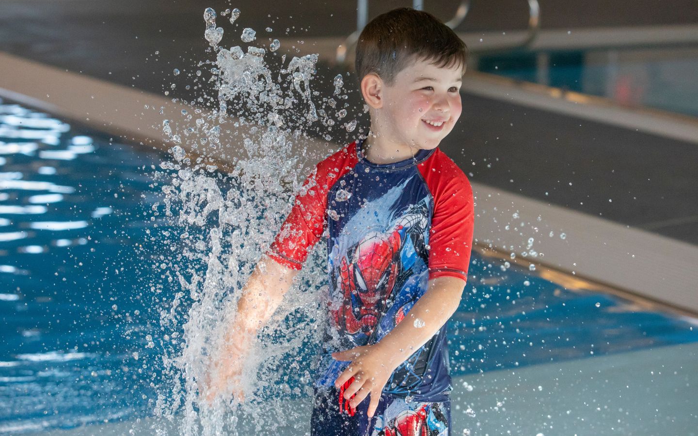 A smiling boy in Spiderman swimmers playing in the toddler pool at the Christchurch Recreation and Sport pool. 