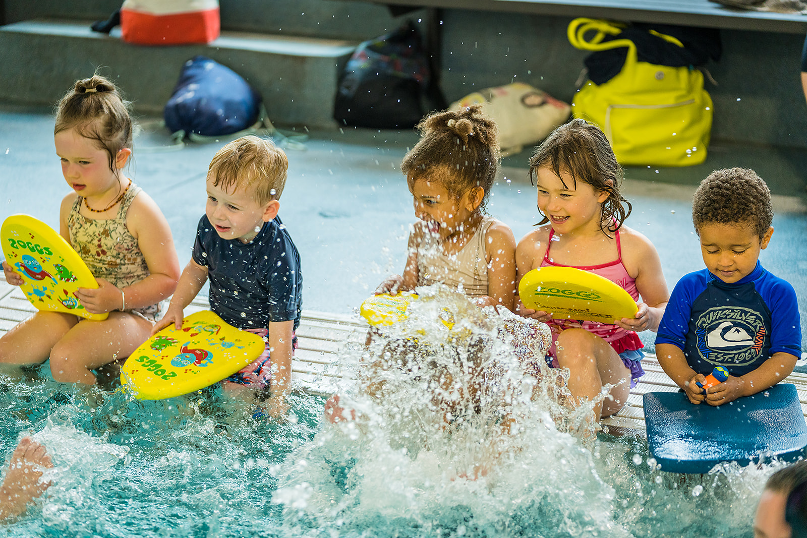 A row of five boys and girls, pre school age smiling and kicking their feet in the water on the pools deck edge holding onto small flotation boards. 